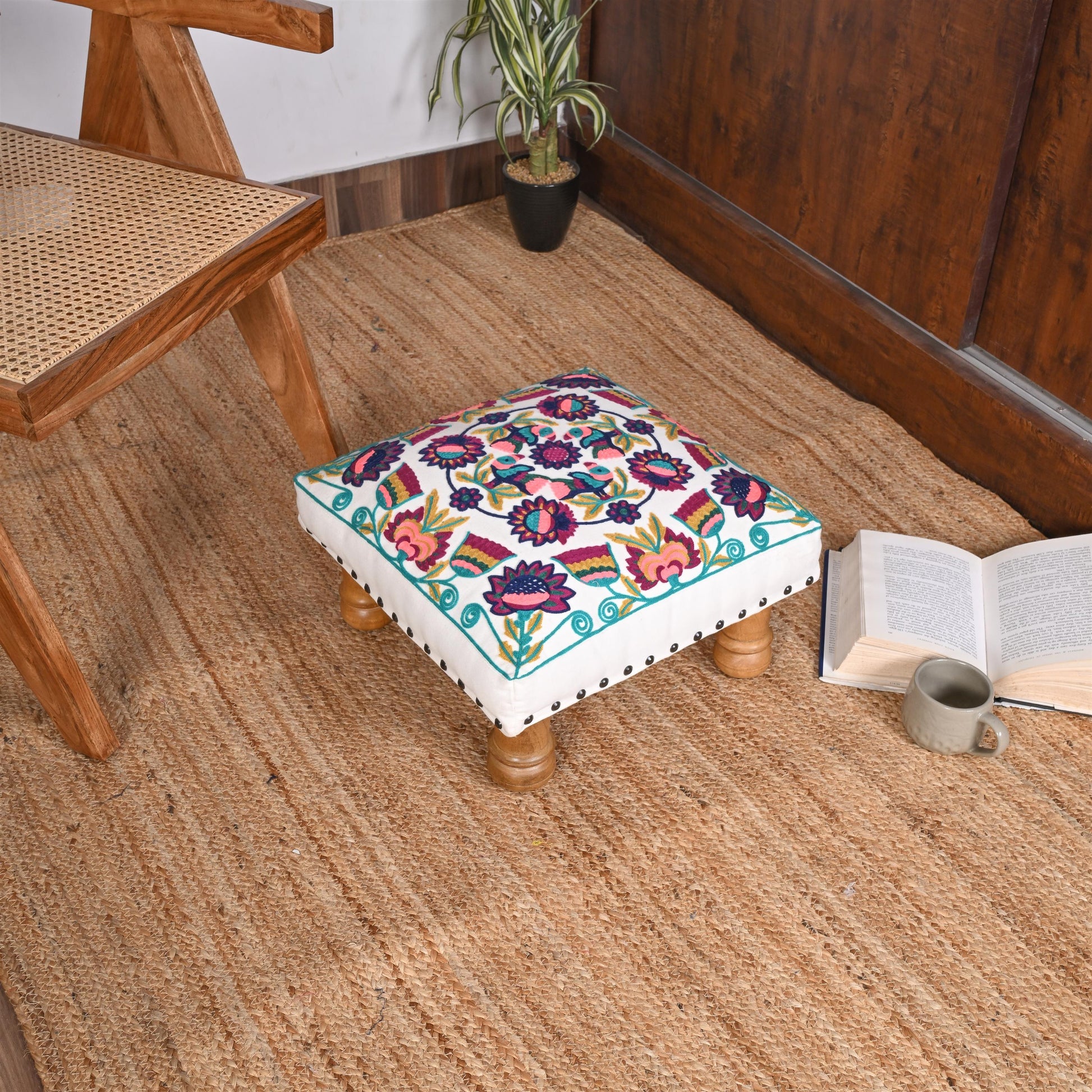 Decorative footstool with floral pattern on a wooden floor next to a chair and open book.
