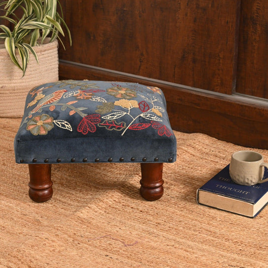 Small ottoman with floral pattern on a wooden floor next to a book and mug.