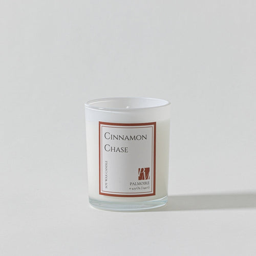 A white glass candle labeled "Cinnamon Chase" with a brown border.