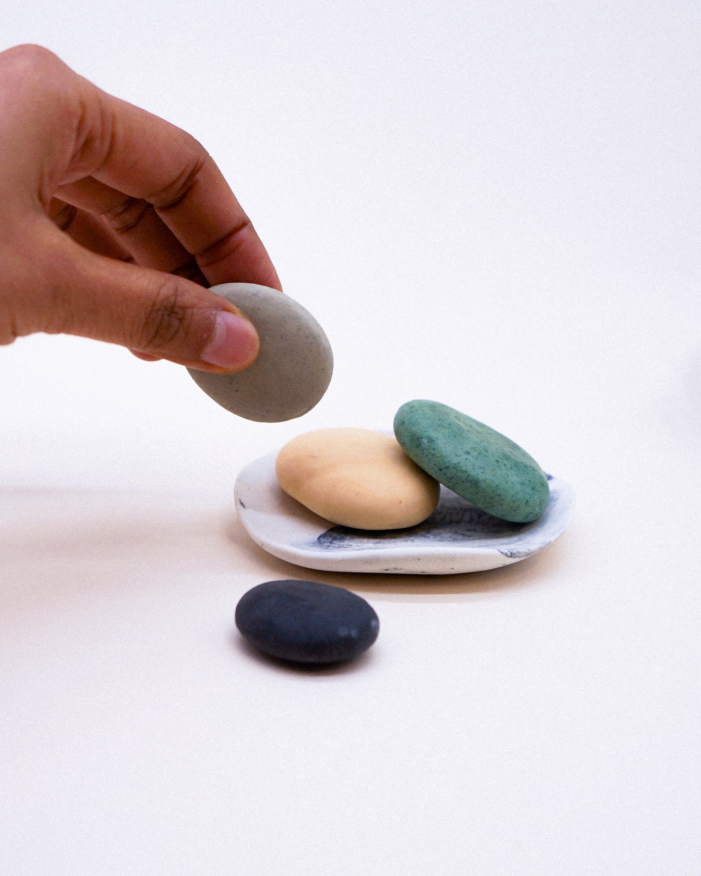 Hand holding a gray stone over a white plate with three painted stones on a white background