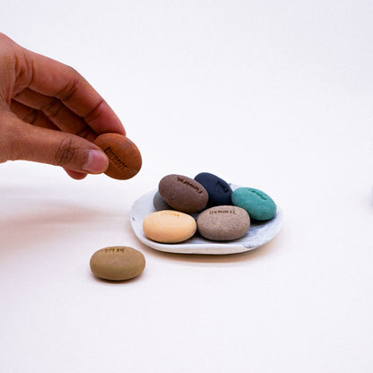Hand holding a brown stone with other stones of different colors on a white plate against a white background