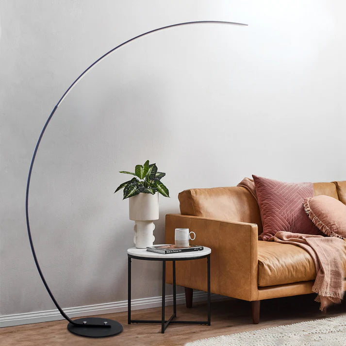Modern black metal curved LED floor lamp arching over a tan leather sofa in a living room.